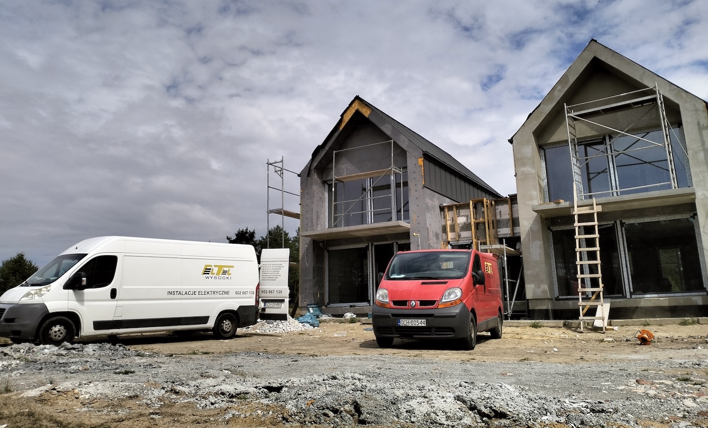 Dwa niedokończone nowoczesne domy w budowie są pokazane z rusztowaniami. Biała i czerwona furgonetka są zaparkowane z przodu na żwirowym podłożu pod częściowo zachmurzonym niebem.