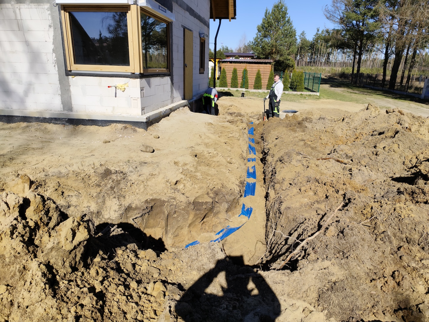 Rów wykopany w ziemi obok domu z niebieską rurą lub kablem ułożonym wewnątrz; dwie osoby stoją w pobliżu sprzętu budowlanego w tle.