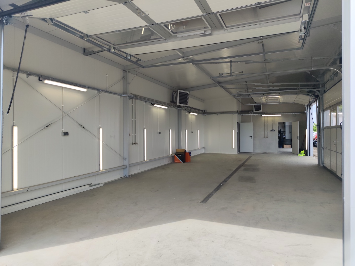 Duży, pusty garaż przemysłowy z białymi ścianami, górnym oświetleniem i otwartymi drzwiami garażowymi wpuszczającymi światło dzienne. Wzdłuż ścian widoczne są narzędzia i sprzęt.