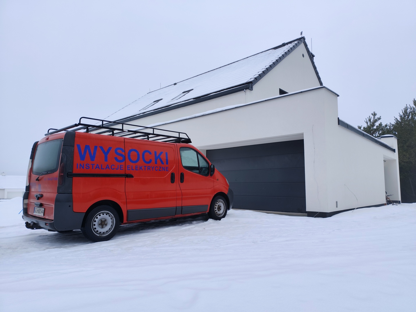 Czerwona furgonetka z napisem "Wysocki Instalacje Elektryczne" jest zaparkowana na zaśnieżonym podjeździe obok nowoczesnego białego domu z garażem.