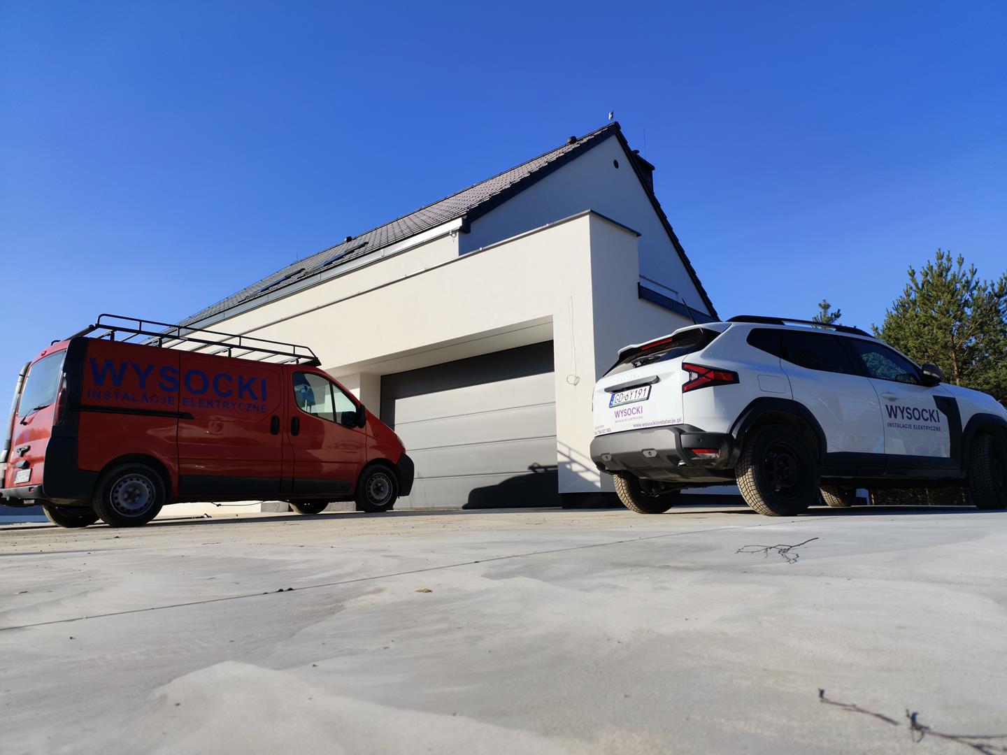 Nowoczesny biały dom z zamkniętym garażem; czerwona furgonetka i biały SUV, oba z napisem "Wysocki", zaparkowane na podjeździe pod czystym błękitnym niebem.